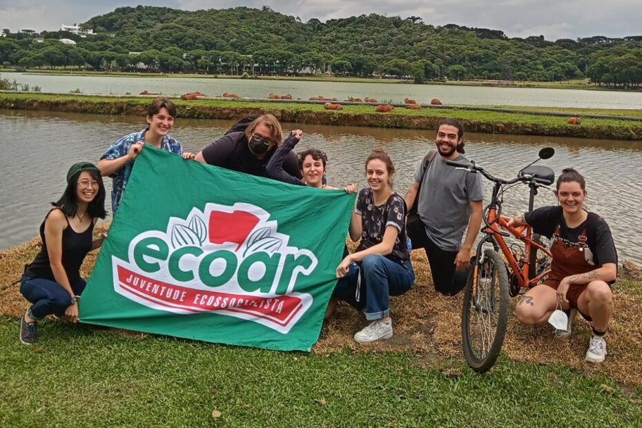 Imagem retangular em formato paisagem. Na foto, que é colorida, se veem sete militantes. Os cinco da esquerda seguram uma bandeira verde com a logo do coletivo Ecoar. A militante mais à direita segura uma bicicleta. Todos estão abaixados na grama. Ao fundo, se veem duas lagoas e diversas capivaras.